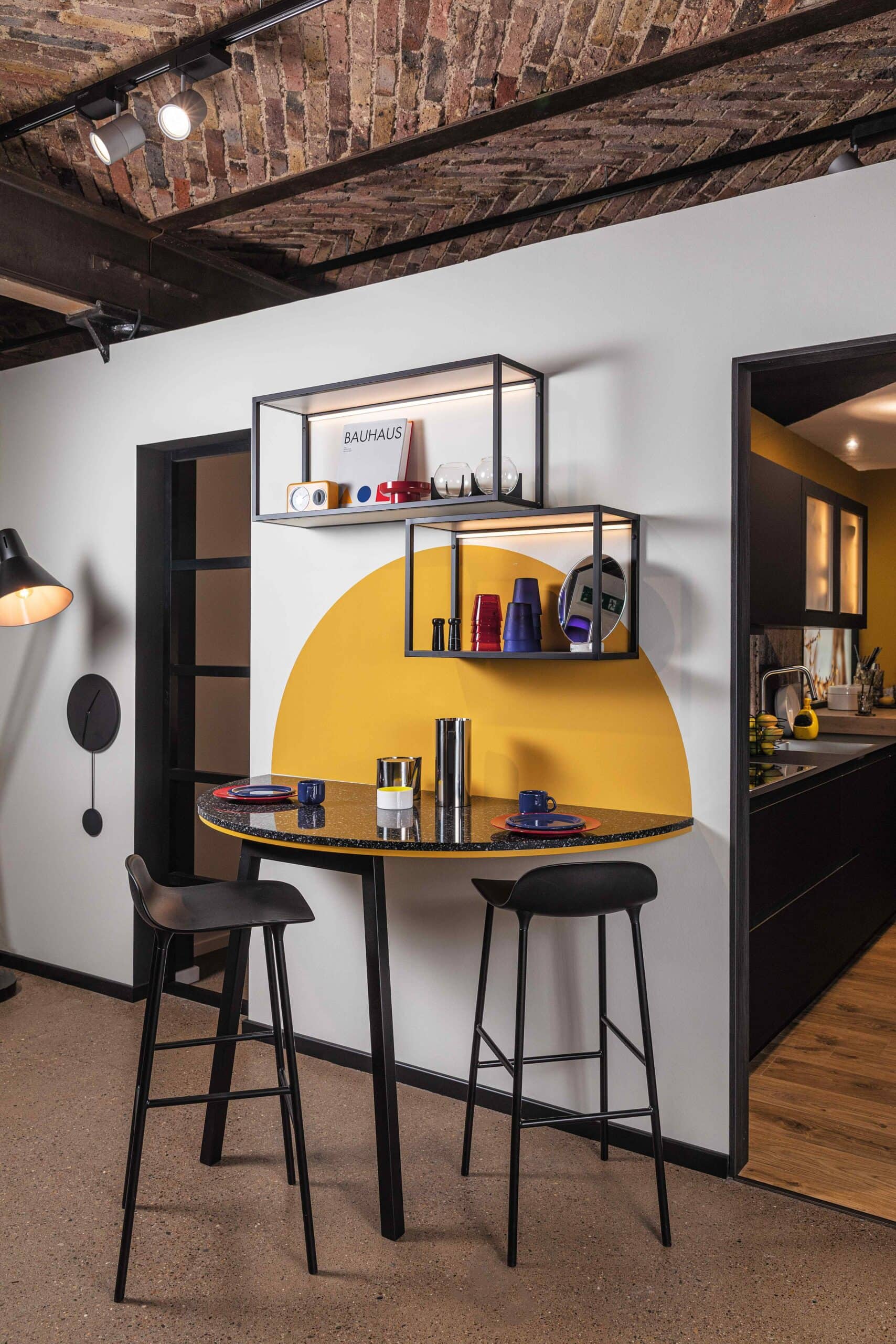 A modern kitchen with a small round table, two bar stools, and a yellow painted semicircle on the wall beneath open shelves holding various items. The space features an exposed brick ceiling and black accents, showcasing innovative solutions for small living rooms integrated into the design.