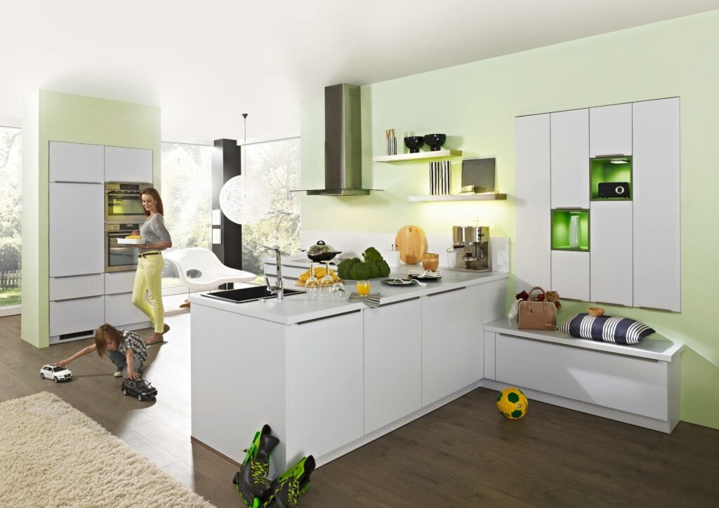 A modern kitchen with white cabinets, an island, and stainless steel appliances, illuminated by sleek German lighting design trends. A woman is using the oven while a child plays with a toy car on the floor. A soccer ball and shoes are nearby.