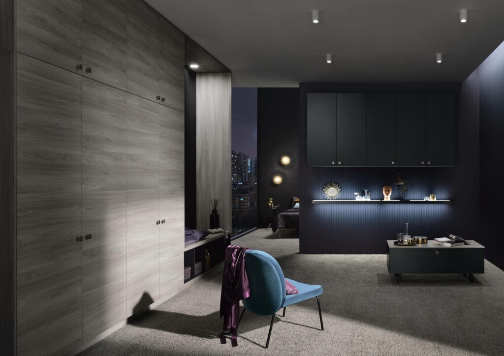 Modern bedroom with a blue chair, wooden cabinetry, and illuminated display shelf inspired by German lighting design trends. A window reveals a cityscape at night. Neutral-toned carpet covers the floor. Modern bedroom with dark blue and wooden tones, featuring a large wardrobe, a chair with draped purple fabric, a low cabinet, and a cityscape visible through the window at night. Adjacent is an open-concept kitchen design seamlessly connecting to a dining area with black chairs in the foreground.