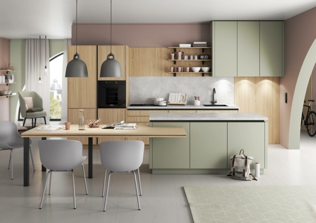 Modern kitchen with light green cabinets, wooden accents, and a grey countertop. Pendant lights hang over a wooden dining table with grey chairs. The room features a minimalist kitchen design enhanced by natural light and an earthy color palette.