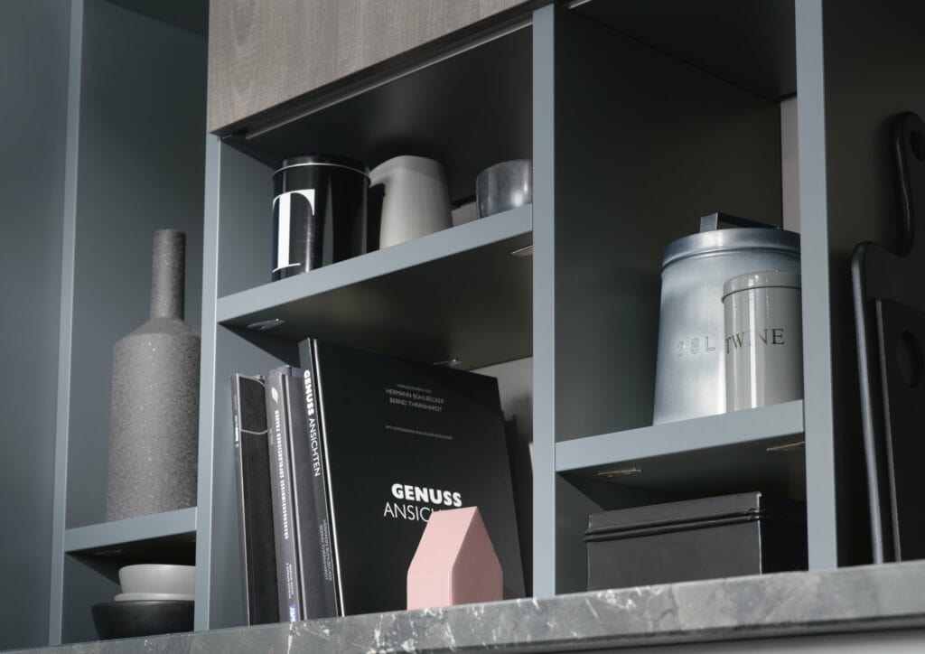 A kitchen shelf with gray ceramic and metal containers, various books, a pink vase arranged in compartments, and a book on the history of German kitchen cabinets.