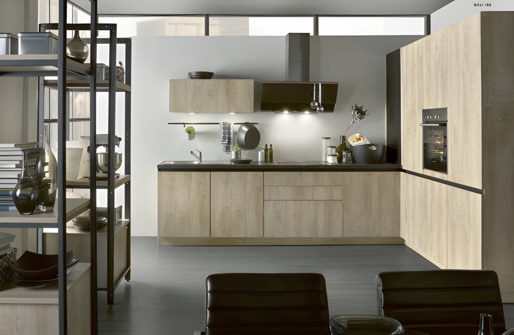 Modern kitchen with light wood cabinets, black countertops, and stainless steel appliances. Shelves with kitchen items on the left, and a wall oven on the right. Two black chairs in the foreground.