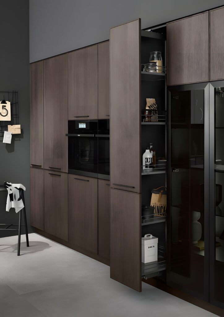A modern kitchen with dark wood cabinets, a built-in oven, and an open pantry shelf displaying various items. The adjacent walls are painted gray. Maximizing storage with pullouts and roll-out shelves, a stool and some decorative items are visible.