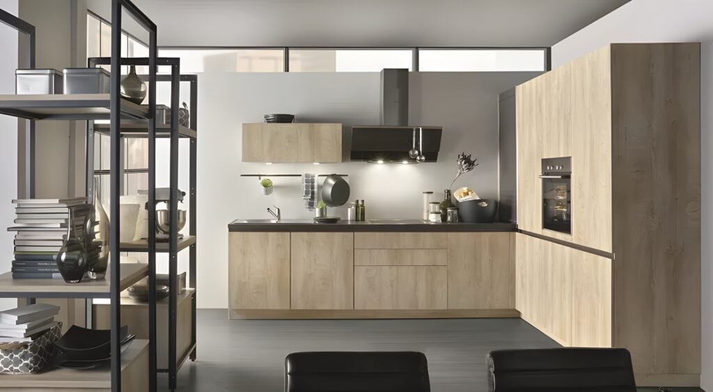 Modern kitchen with light wood cabinets, black countertops, built-in oven, sink, stove, and a shelving unit on the left holding books and decorative items. A spotlight as part of the German kitchen lighting design trends highlights the elegant details.
