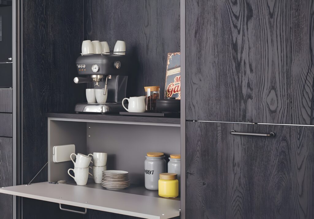 A black coffee machine with cups on top is placed on a shelf integrated into black wood cabinetry, reflecting healthy kitchen design trends. Below it, there are cups, saucers, jars, and a canister on another shelf within the pantry. The cabinet door is open.