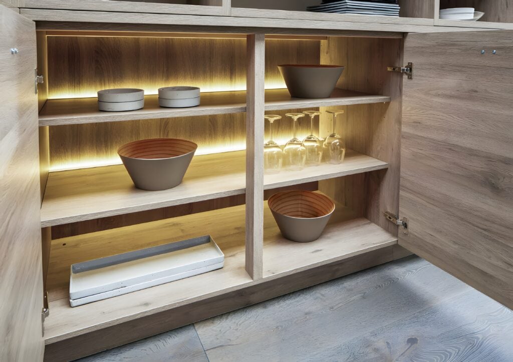 Open wooden cabinet with LED lighting, showcasing bowls, plates, glasses, and a rectangular dish on three shelves—a perfect example of kitchen design trends for home cooks.