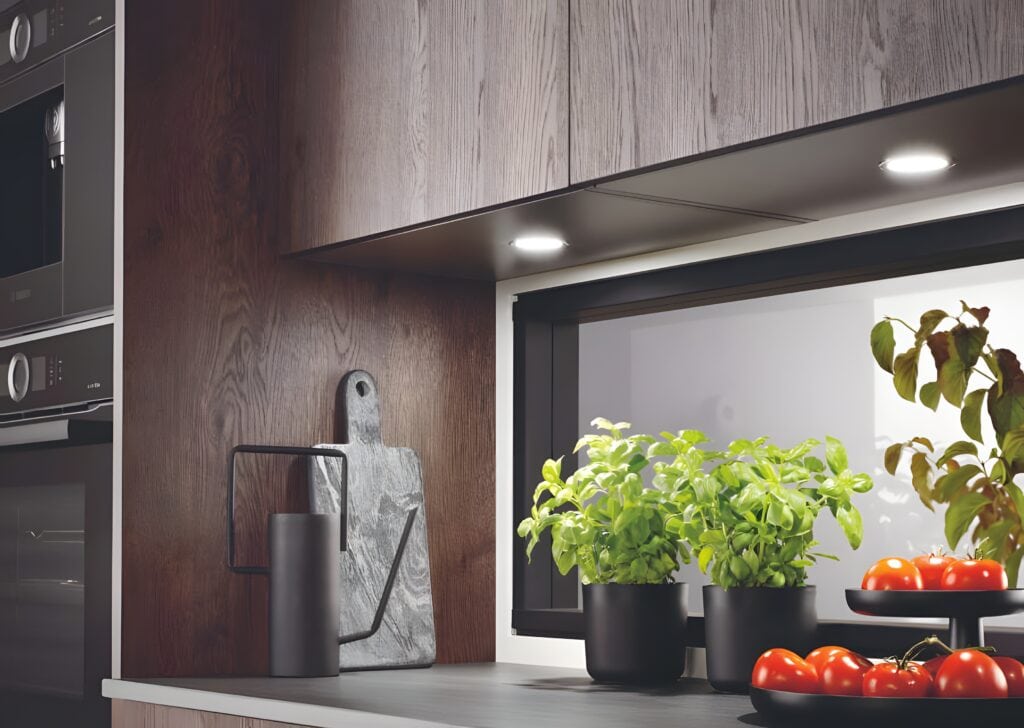 Modern kitchen counter with potted herbs, tomatoes in a bowl, a cutting board, and a sleek metallic jug under warm lighting, reflecting the latest health-focused kitchen design trends.