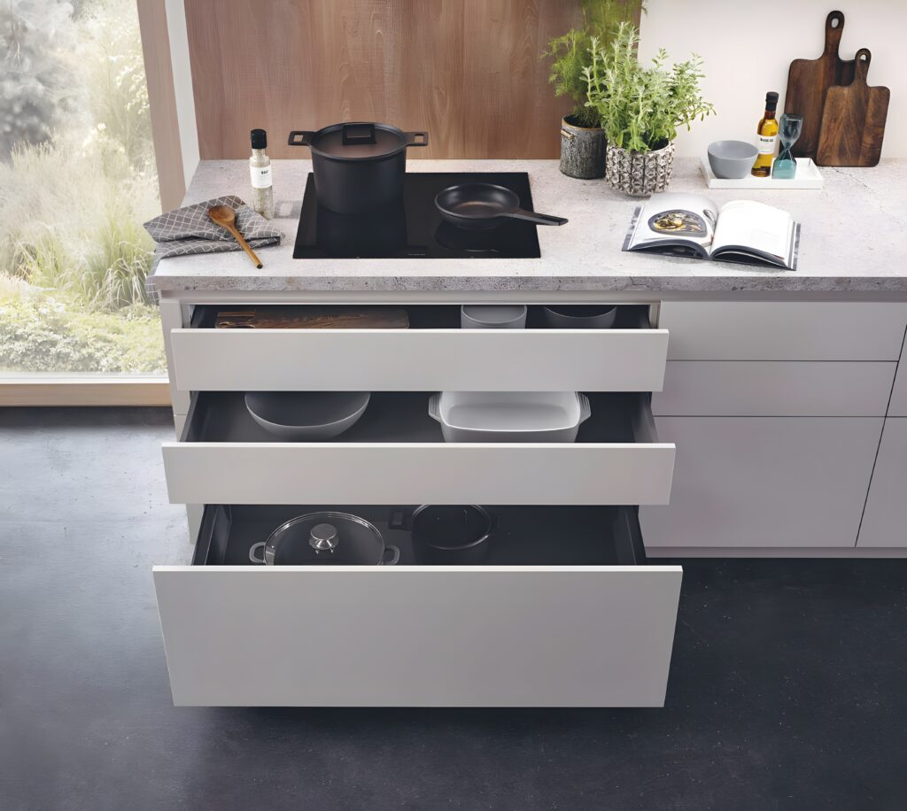 Modern kitchen with an induction cooktop, pot, and pan on the counter. Three open drawers reveal innovative storage solutions in German kitchens for utensils and cookware. An open cookbook, cutting boards, spices, and a plant are also visible.