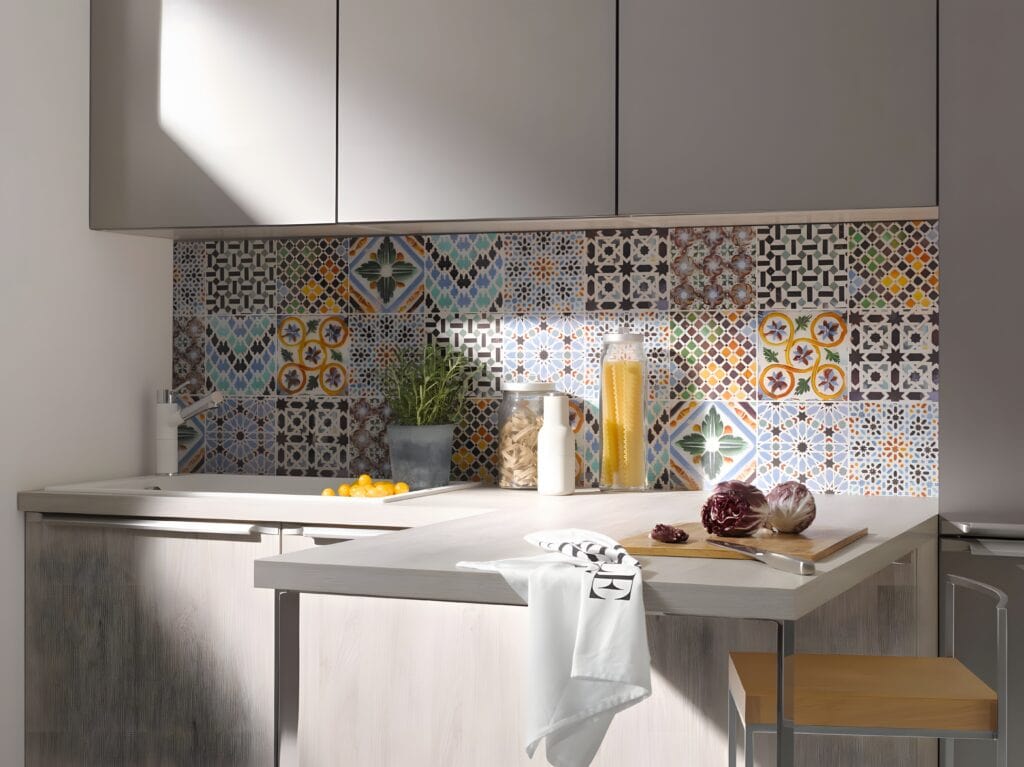 A modern kitchen with a colorful tiled backsplash, light gray cabinets reminiscent of the history of German kitchen cabinets, a countertop with various items including a cutting board, vegetables, and a white towel hanging from the counter edge.