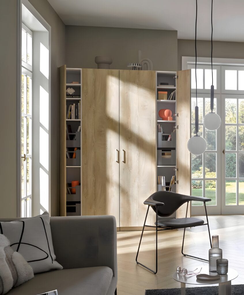 Modern living room with a large wooden cabinet, a grey armchair, and minimalist decor. Sunlight streams through tall windows, casting a bright atmosphere throughout the space.