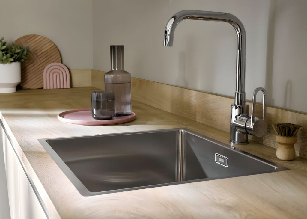 A modern kitchen sink with a stainless steel faucet sits in a light wooden countertop, reflecting the latest kitchen design trends for home cooks. A tray with glass and ceramic items, along with a potted plant, is placed to the left of the sink.