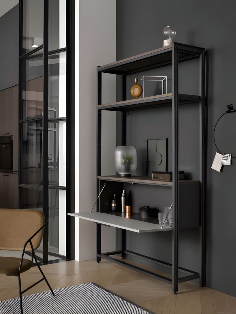 Introducing the modern shelving unit with Creative Storage and Organization by Bauformat. It includes various items like a glass vase, framed artwork, and a light bulb, all on display. Featuring an integrated desk space, it stands against a dark wall and elegant glass partitions. A modern, minimalist shelving unit with various decor items and a fold-out desk sits against a dark wall. The space-saving kitchen furniture is complemented by a beige chair and a grey rug in the foreground.
