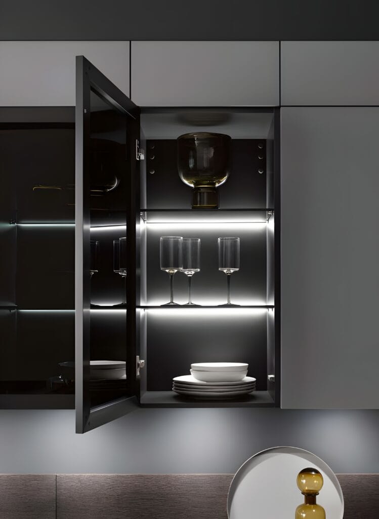 Open kitchen cabinet with interior lighting reveals glass shelves holding several drinking glasses, a bowl stack, and a large container—modern innovations in German kitchen design at their finest.