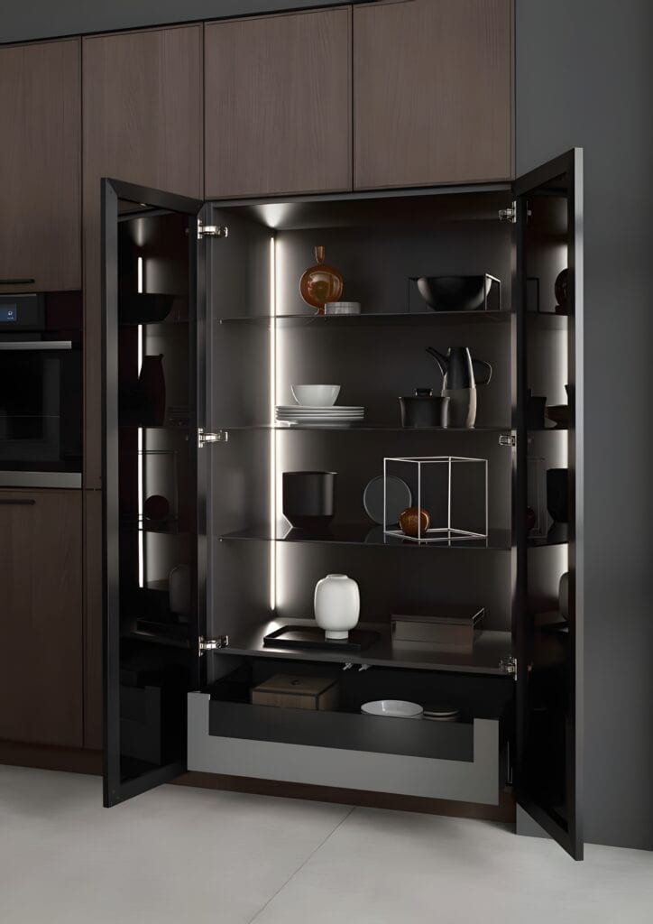 Open kitchen cabinet with black and white dishware, including bowls, plates, and a teapot, neatly arranged on glass shelves. Highlighting key trends in smart kitchen cabinet materials, the unit features interior lighting and a closed drawer at the bottom.