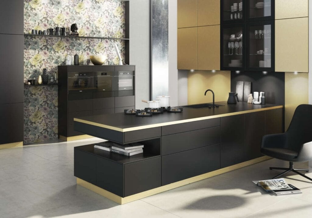 Modern kitchen with black and gold cabinets, a center island with a stovetop, floral backsplash, built-in oven, and wall-mounted shelves. Reflecting healthy kitchen design trends, a black chair is placed on the right side for casual dining or meal prep.