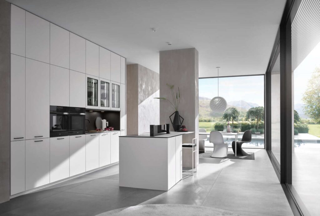 Modern, minimalist kitchen design with white cabinets, a central island, and a dining area. Large floor-to-ceiling windows offer a view of the patio, greenery, and mountains in the background.