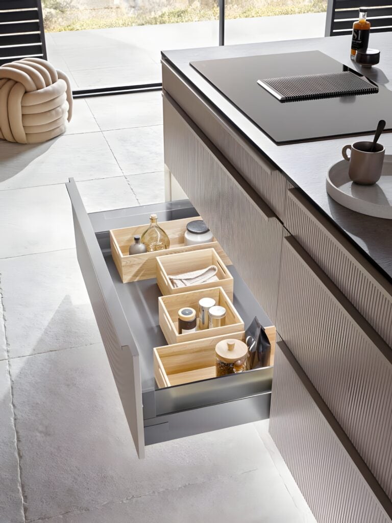 A kitchen drawer is pulled out, revealing custom storage solutions with neatly organized wooden boxes holding various kitchen items. The countertop above boasts a cup, a chopping board, and a bowl, while a window frames an inviting outdoor view.