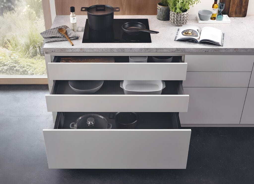 Open kitchen drawers under a sleek marble countertop, revealing innovative storage for pots, pans, and bowls. The minimalist kitchen design features a cookbook, oil bottles, potted plants, and a stove with a pot and pan on the pristine countertop.