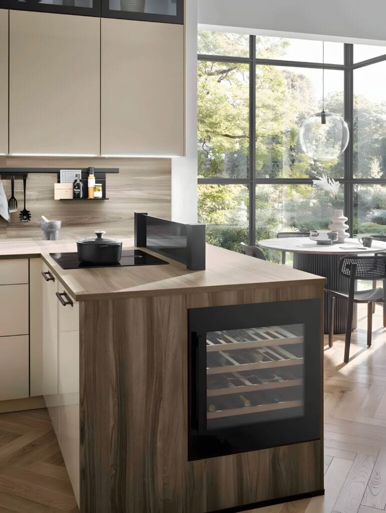 Modern kitchen with beige cabinets made from eco-friendly materials, a wooden island featuring a wine cooler, induction cooktop, and black accents. Large windows in the background open to a dining area with chairs and a round table.
