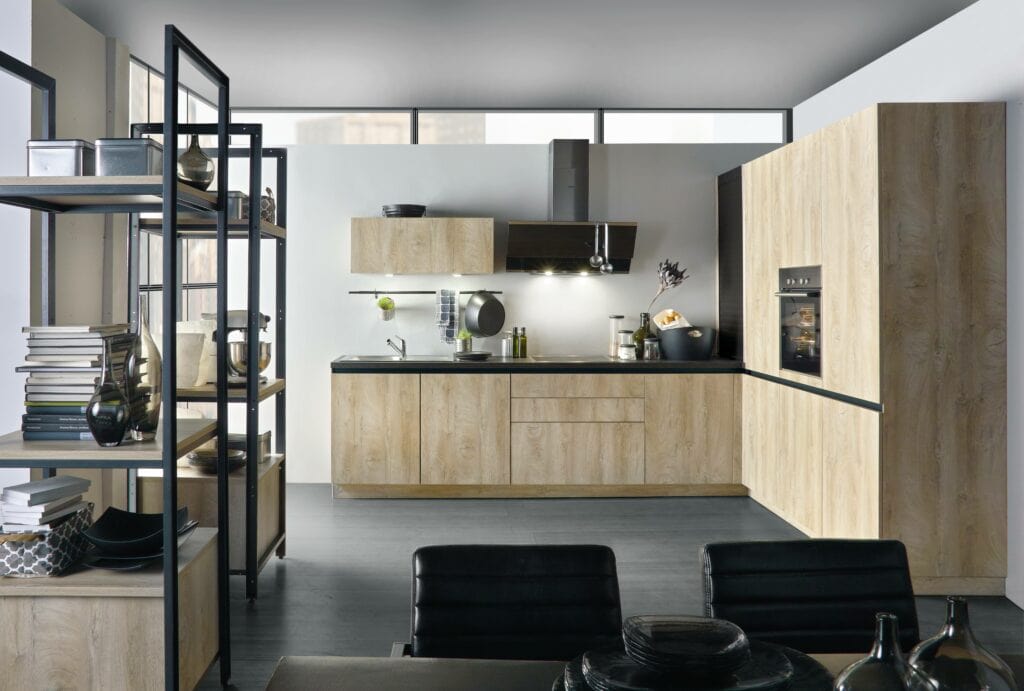 Modern kitchen with light wood cabinets, a black countertop, stainless steel appliances, and open shelving. The open-concept kitchen design seamlessly connects to a dining area with black chairs visible in the foreground.