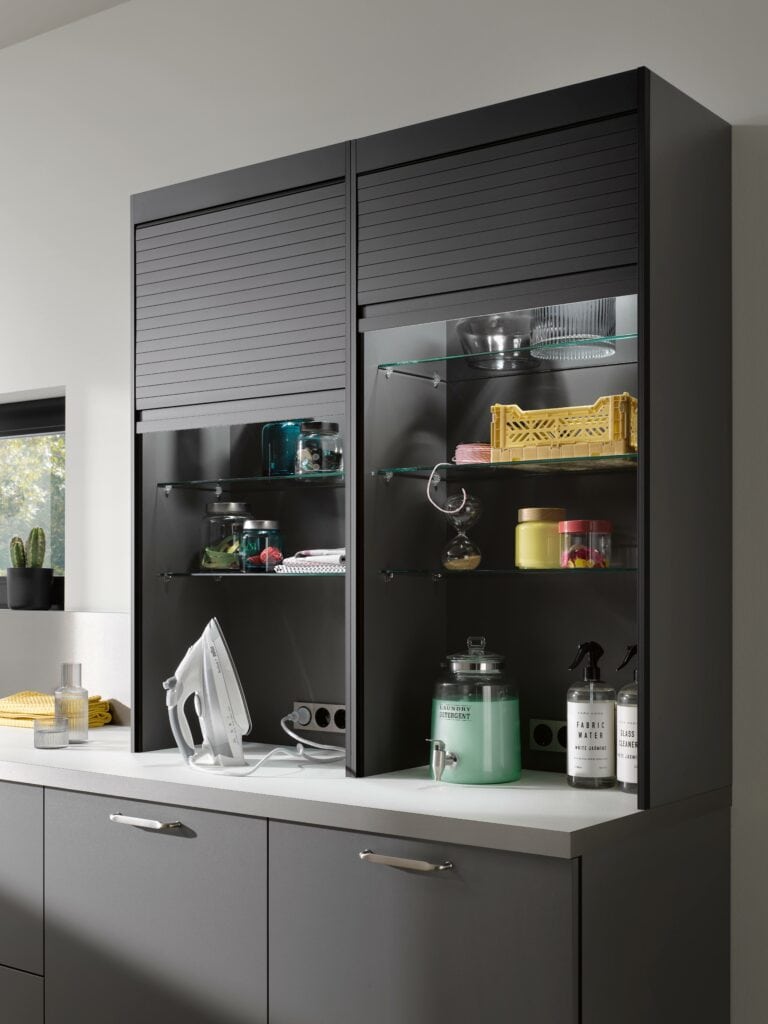 Gray kitchen cabinets with roller shutter doors; one door is open revealing shelves with kitchen items such as pots, containers, and an iron. Countertop has an iron, jars, soap dispenser, and cleaning items. These cabinets highlight the advantages of smart materials in modern kitchen design.