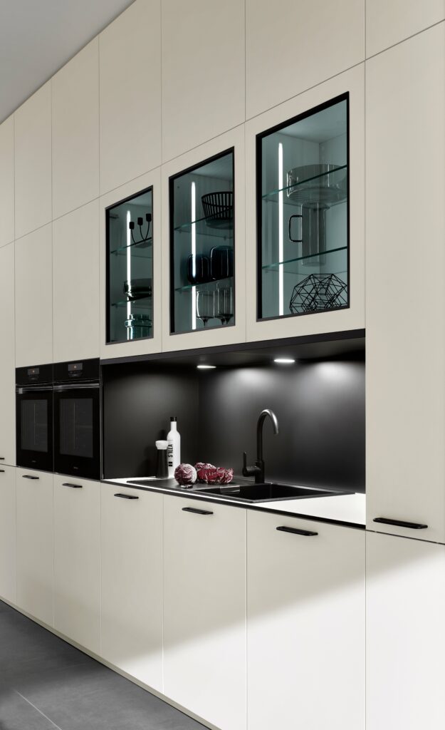 Modern kitchen with white cabinets, black countertop, and a black backsplash. The upper cabinets have glass doors showcasing kitchenware. A sink with a black faucet and some kitchen items are on the counter, reflecting contemporary healthy kitchen design trends.