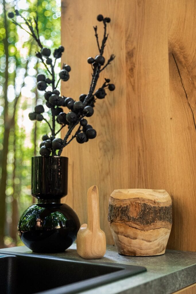 A vase with branches, a decorative wood object, and a wooden container are placed on a countertop near a sink. The setup is beautifully complemented by eco-friendly kitchen cabinets against the wooden wall background.