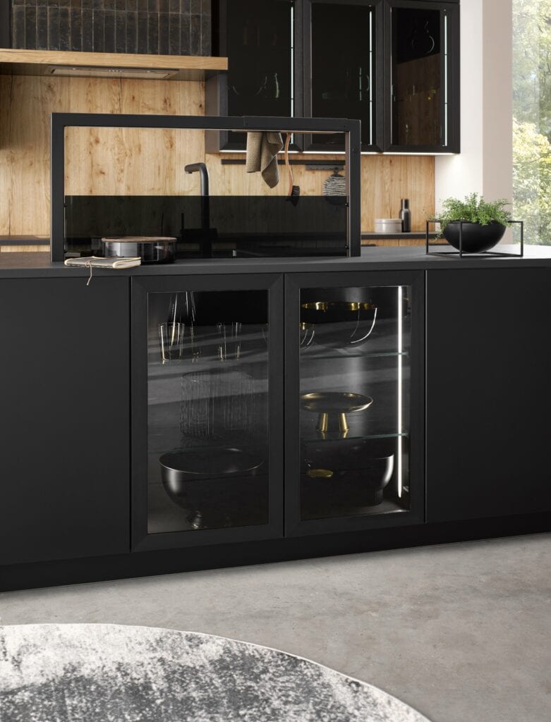 A modern kitchen with black cabinets, a glass-front cabinet displaying kitchenware, and a black countertop. The wooden backsplash and shelf add a contrast to the sleek design, showcasing glass elements in German kitchen design.