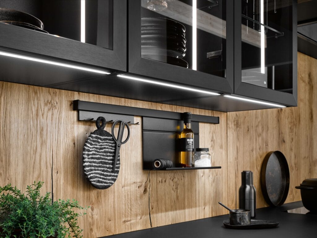 Modern kitchen with black cabinetry, a wooden backsplash, and a black counter. Shelves and hooks hold kitchen items including a hanging scrubber, bottles of oil, a spice jar, and a black pepper mill.