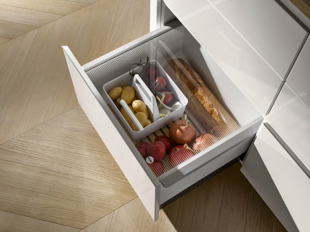 Open kitchen drawer with a baguette, potatoes, tomatoes, and onions, organized in compartments. The drawer is part of a white, modern kitchen unit that highlights the role of engineering in durability.