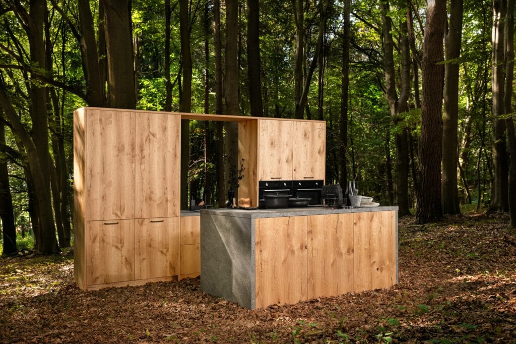 A modern wooden kitchen set with a stone countertop is placed in a forest, surrounded by trees and fallen leaves, embodying the trend toward eco-friendly materials.