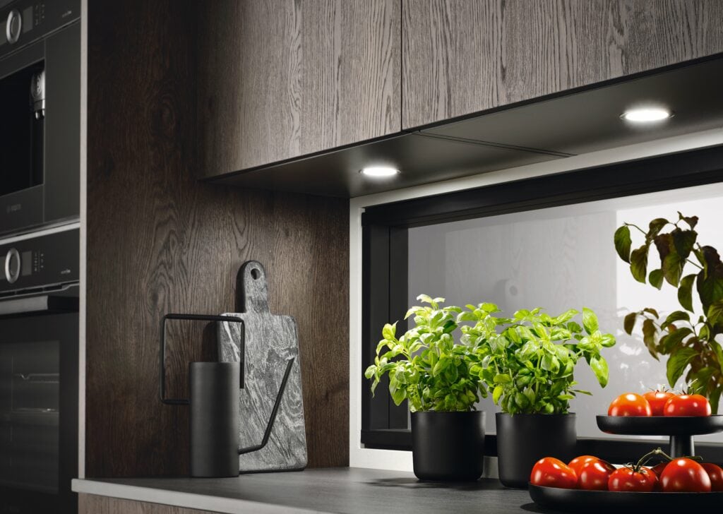 A modern kitchen countertop with a potted basil plant, a black watering can, a wooden cutting board, tomatoes, and leafy greens.
