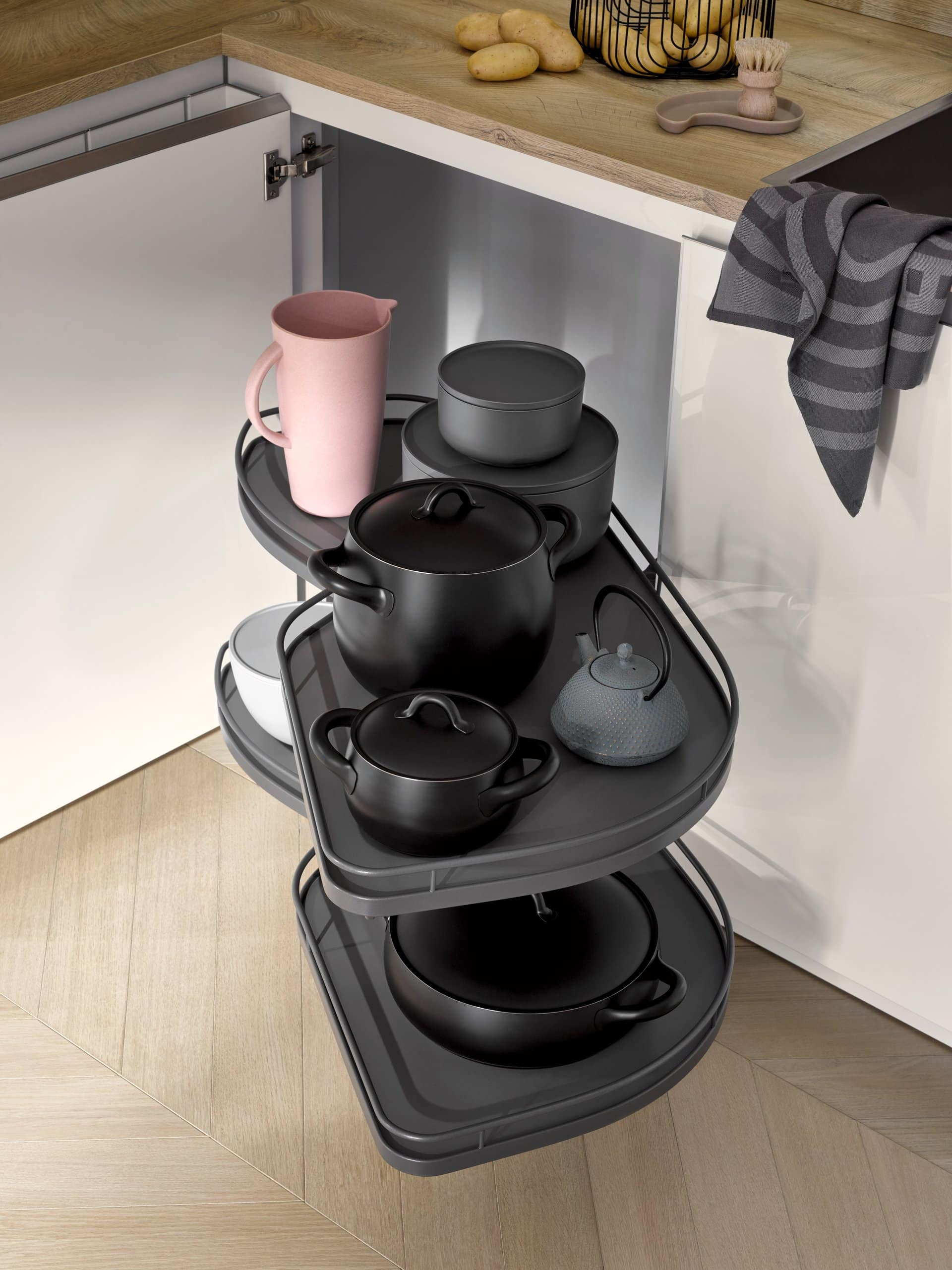 Pull-out corner shelf inside a kitchen cabinet, displaying various black pots, a teapot, bowls, and a pink pitcher. A striped towel hangs over the countertop edge. This space-saving kitchen furniture maximizes storage while keeping essentials within easy reach.