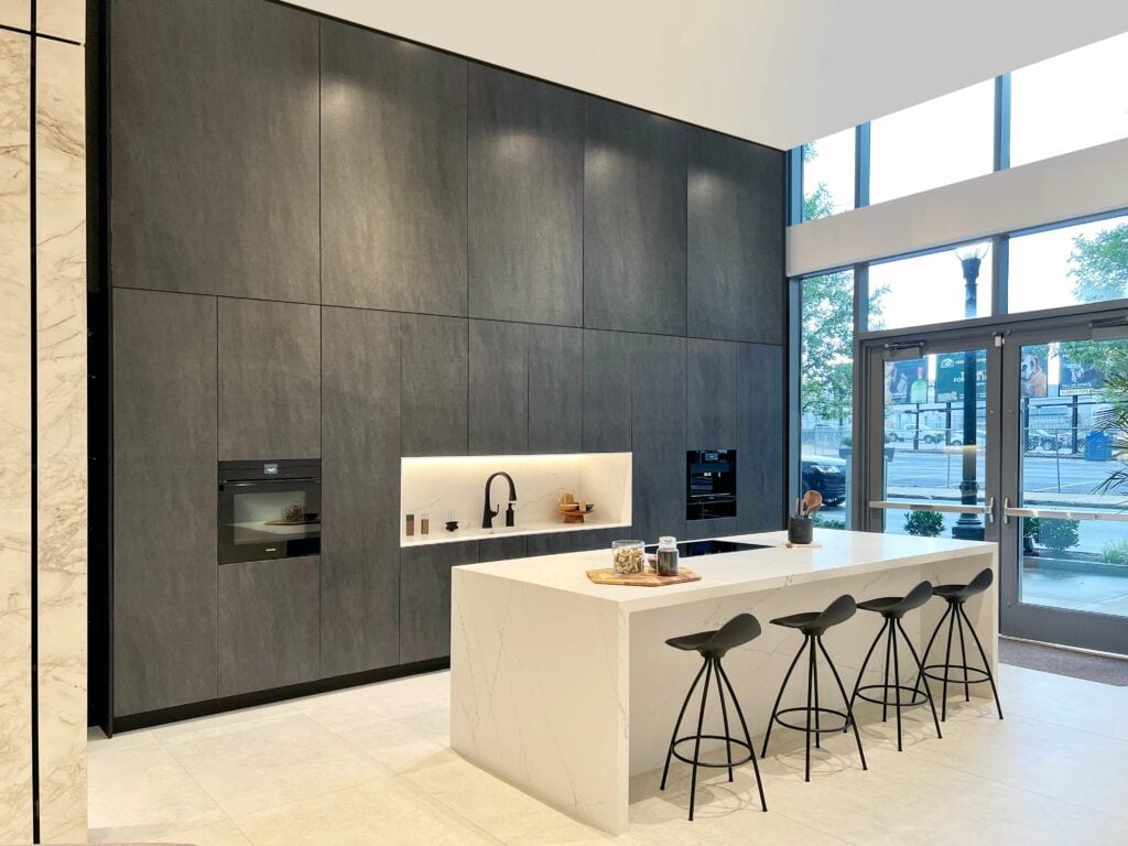 Modern, open concept kitchen design with dark cabinets, built-in appliances, and a white island with sink and bar stools. Floor-to-ceiling windows flood the space with natural light, and a striking marble wall graces the left side.