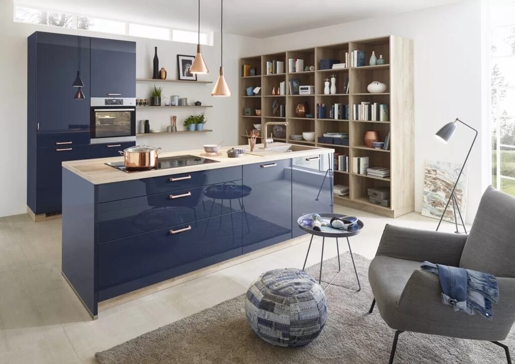 Modern kitchen and living area with clean and care high gloss navy cabinets, a wooden kitchen island, open shelving with books and decor, a gray armchair, and a pouf. Natural light fills the space through large windows.