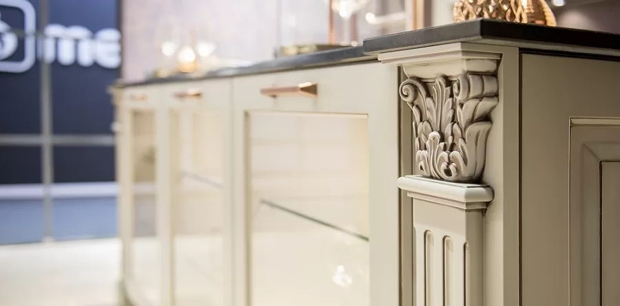 A close-up of an ornate kitchen island featuring intricate carved details on a cream-colored corner pillar, with a black countertop and brass handles on the drawers, reminiscent of the artistry seen in traditional German kitchen cabinets.