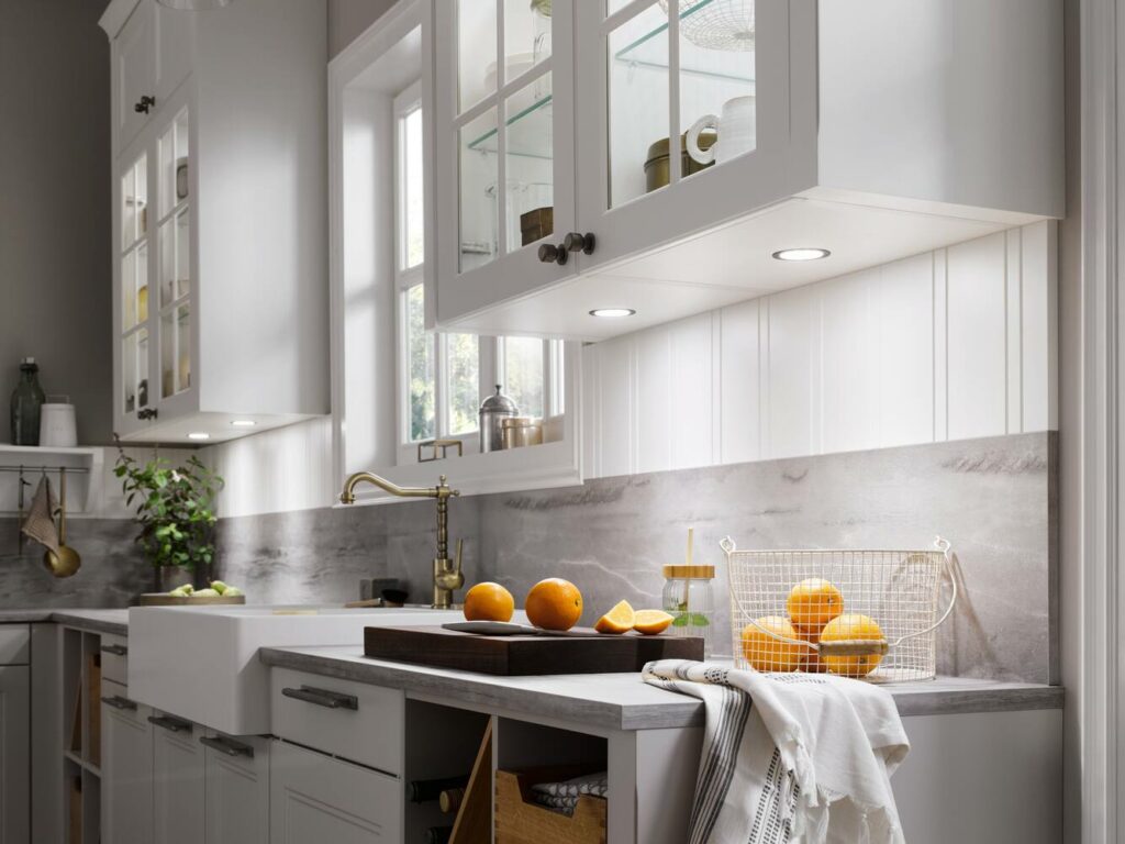 Modern kitchen with white cabinets, marble countertops, and a basket of oranges. Under-cabinet lights illuminate the backsplash. A sink with a brass faucet and a window are in the background, showcasing healthy kitchen design trends.