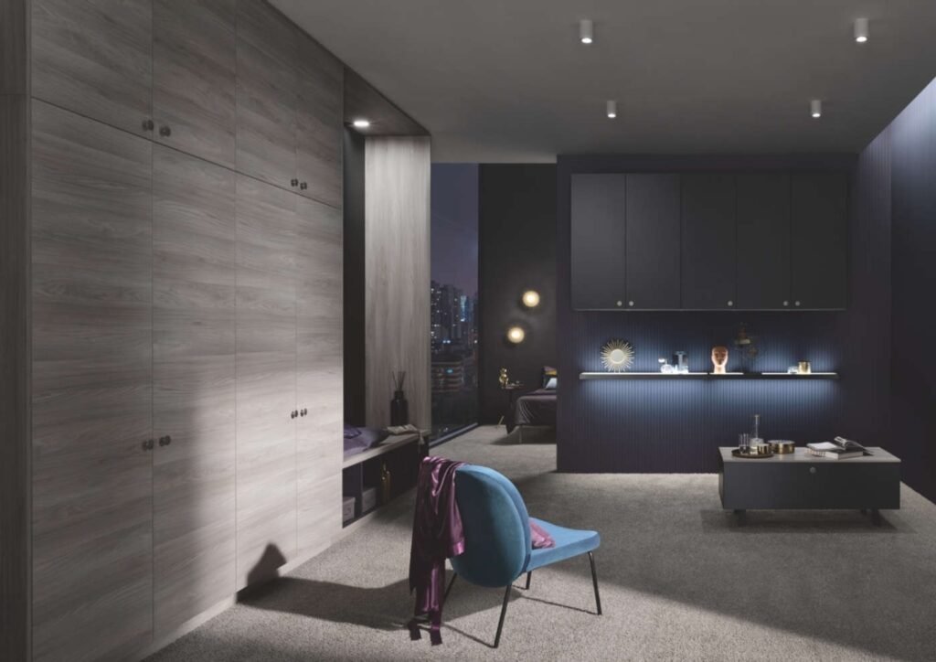 Modern room interior with wooden cabinets, a blue accent wall, a blue chair, and various decor items on a shelf and table. The room is dimly lit with spotlights inspired by German kitchen lighting design trends and has a night cityscape view.