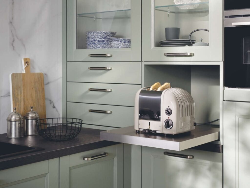 A modern kitchen with green cabinets, a wooden cutting board, a wire basket, and a stainless steel toaster on an extended counter toasting two slices of bread showcases modern innovations in German kitchen design.