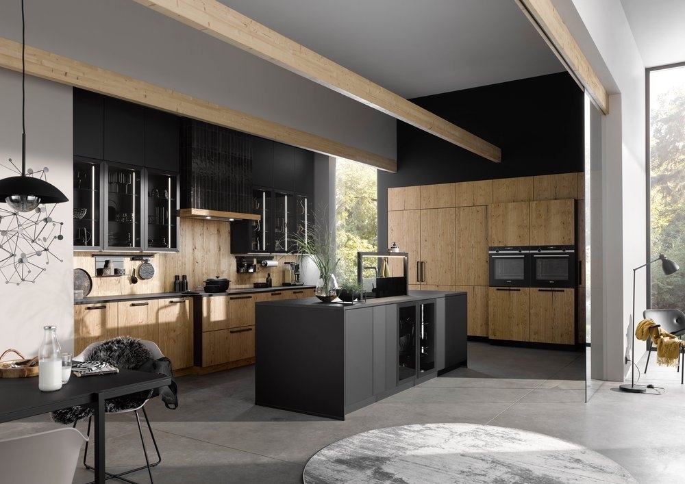 A modern kitchen unearthing Germany's trendsetting kitchen designs, featuring wooden and black cabinets, a large island counter, and minimalist decor. A dining area is visible on the left, while large windows flood the space with natural light.